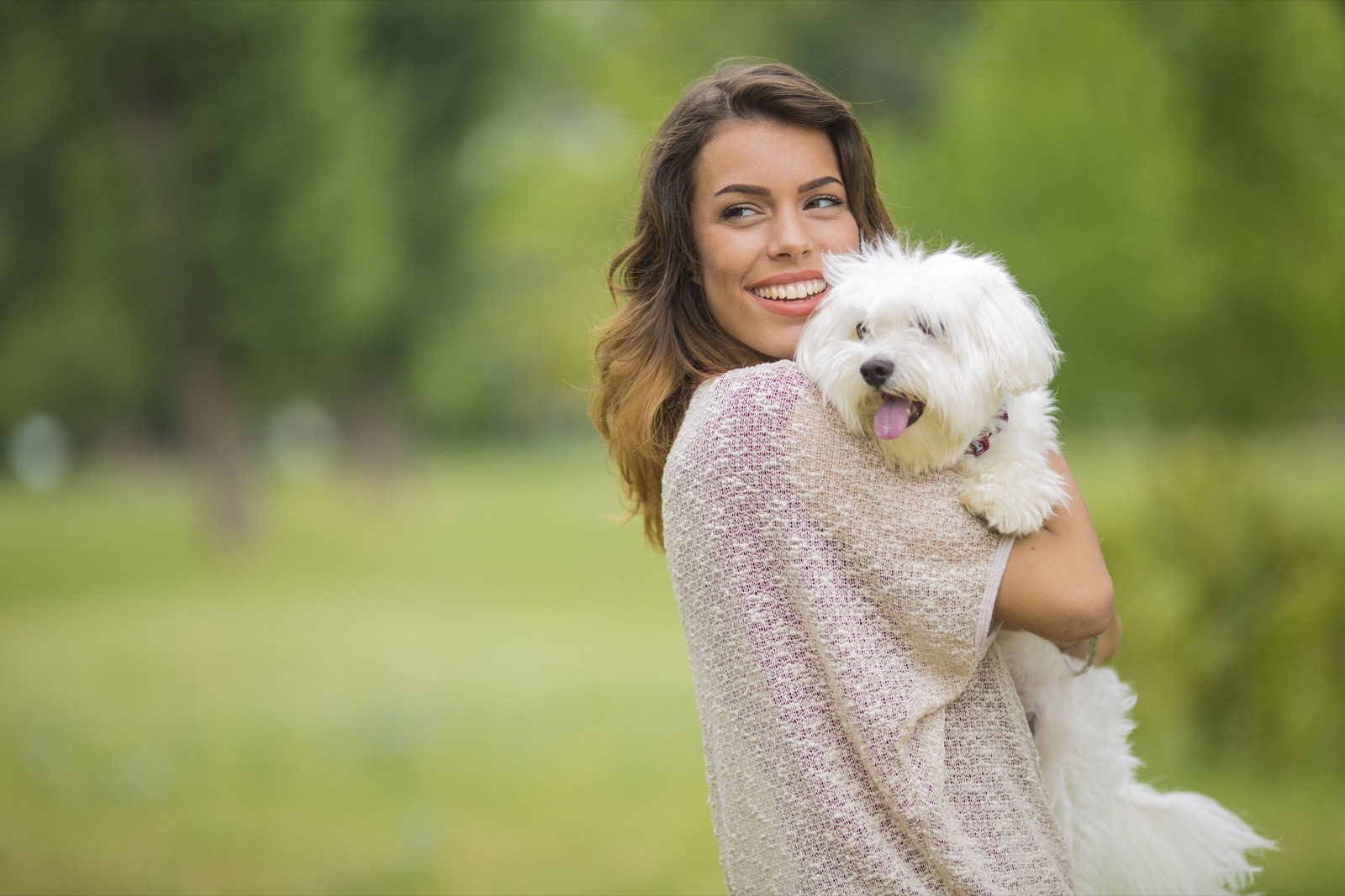 A smiling woman holding a small white Maltese dog in a sunny park.